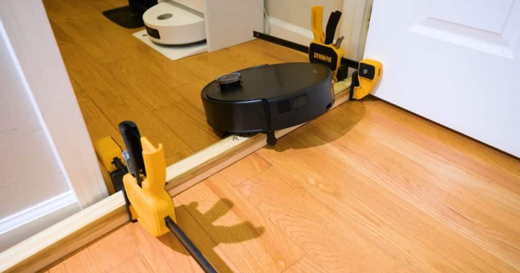 MOVA Z60 Ultra Roller robot vacuum climbing a wooden threshold barrier that's been secured with yellow clamps during testing, evaluating its StepMaster 2.0 lifting system on hardwood floors.