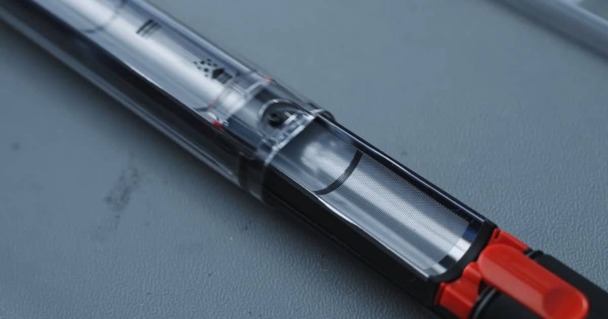 | Vacuum Wars Close-up of the Dyson PencilVac dustbin showing a slim transparent bin with a metal mesh filter and a red latch, highlighting the internal debris-compaction mechanism.