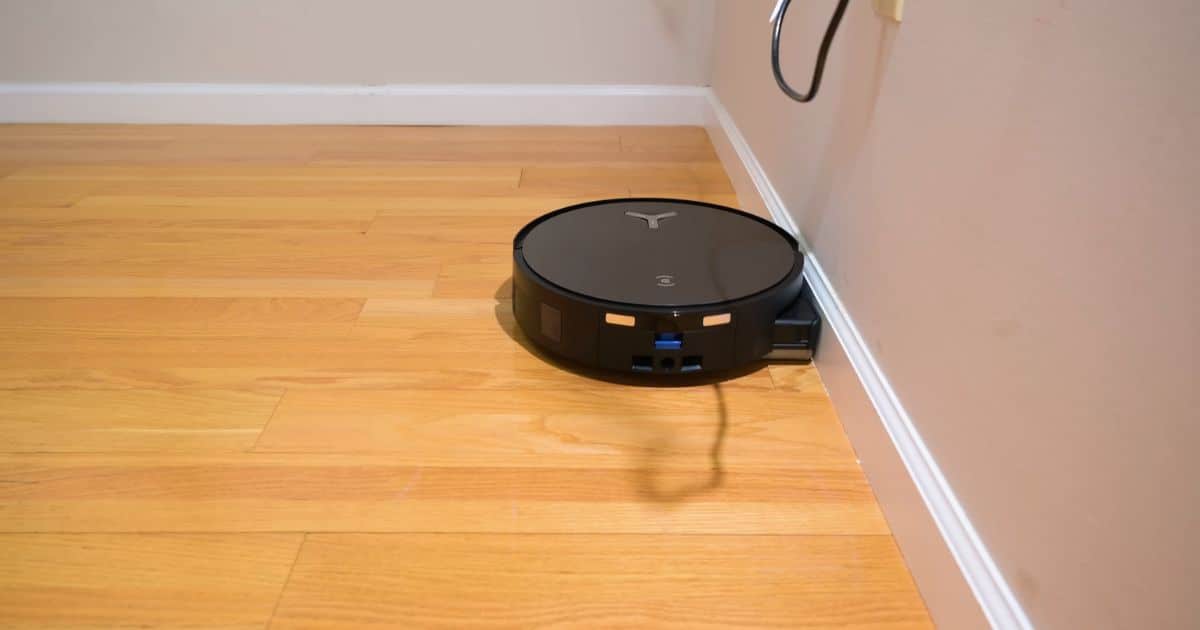 | Vacuum Wars A black robot vacuum with a single roller mop drives along a baseboard on a hardwood floor, positioned tightly to the wall for edge cleaning.