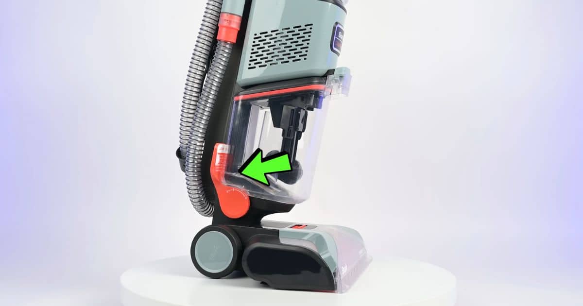 | Vacuum Wars Close-up of a Shark CarpetXpert upright with an onboard hose; a green arrow points to the red/orange physical switch near the base used to route suction between hose cleaning and floor-head cleaning.