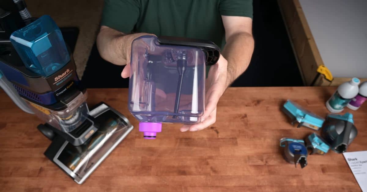 | Vacuum Wars Person holding a removable translucent water tank with a purple cap above a workbench, with a Shark CarpetXpert upright and several attachments and solution bottles nearby.