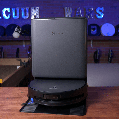 Ecovacs T90 Pro Omni in studio 450 ©Vacuum Wars | Vacuum Wars Robot vacuum docked on its charging base on a wooden table, with a blue neon 'VACUUM WARS' sign in the background.