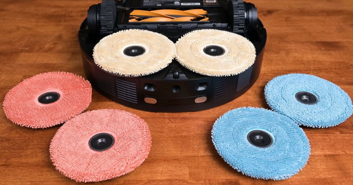 | Vacuum Wars Close-up of the Mobius 60 underside with two beige spinning mop pads installed. Additional red and blue circular mop pads are laid out on a wooden surface in front of the robot.