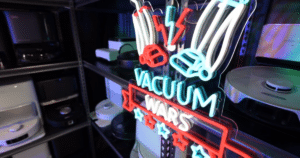 Recent Changes at Vacuum Wars | Vacuum Wars Vacuum Wars neon logo in front of a storage shelf contraining robot vacuums they have purchased and evaluated