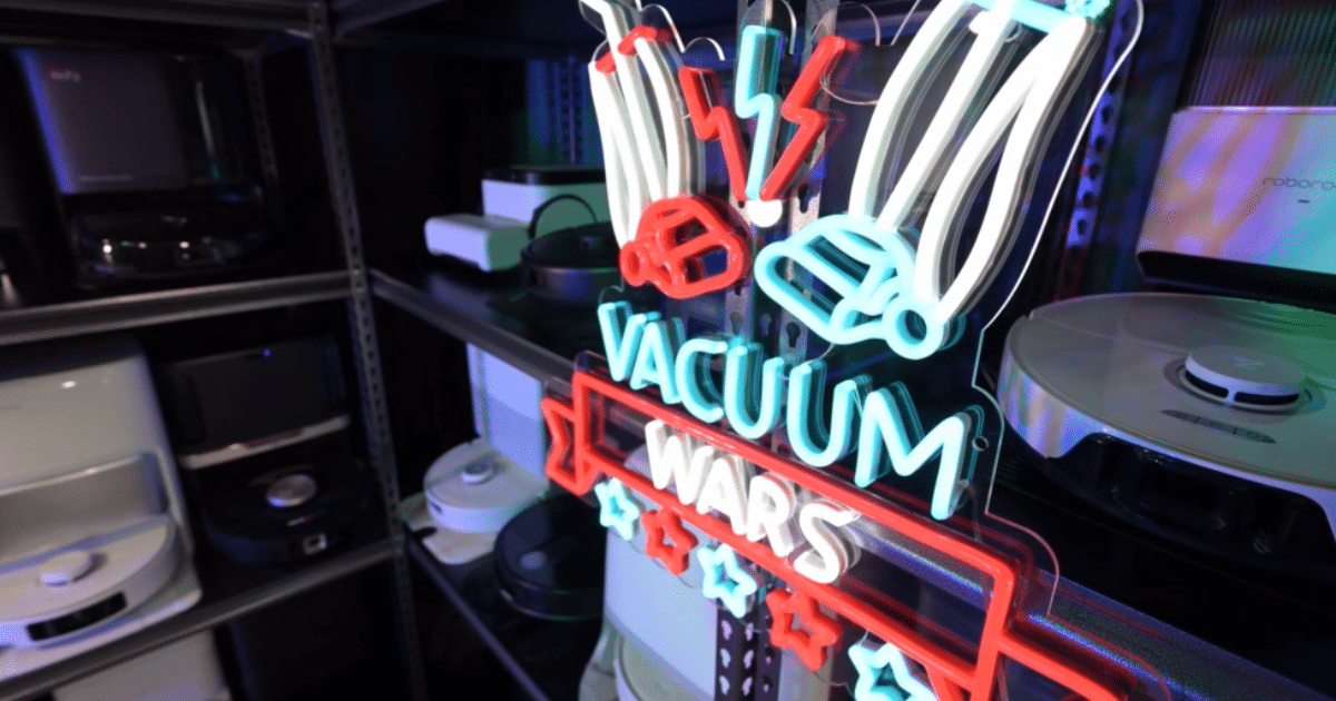 Vacuum Wars neon logo in front of a storage shelf contraining robot vacuums they have purchased and evaluated