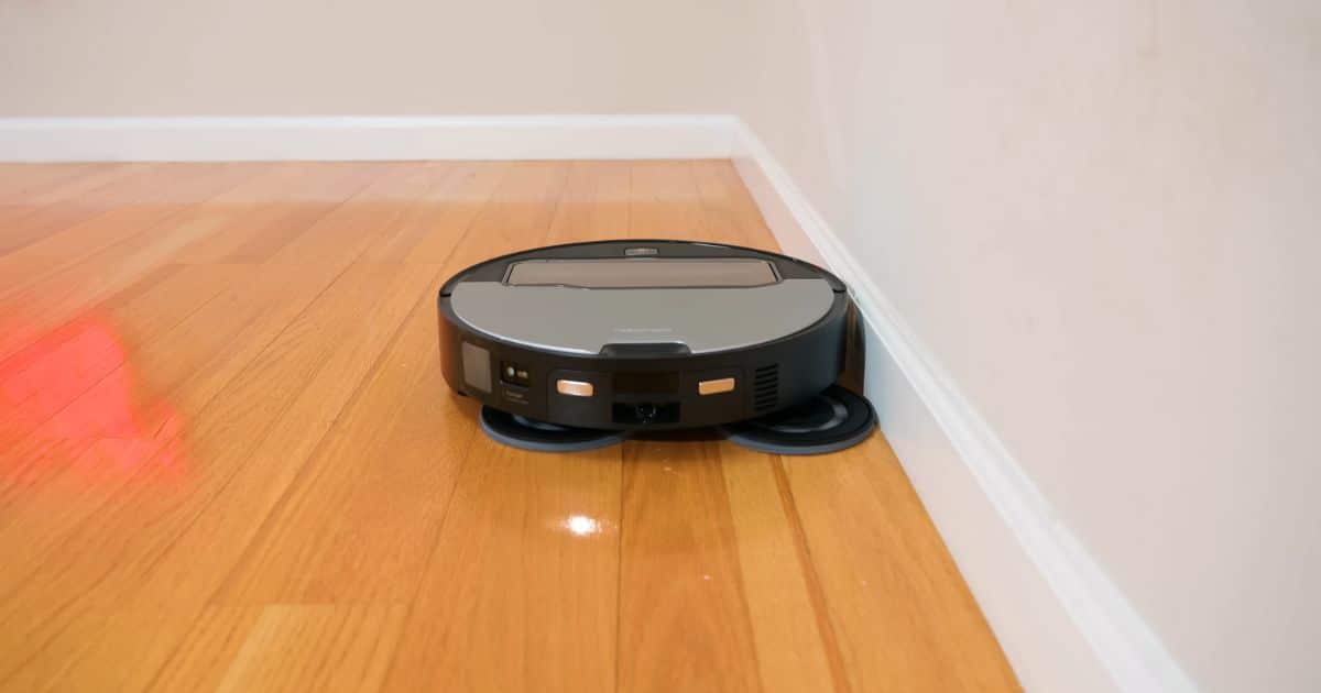 | Vacuum Wars A robot vacuum with dual spinning mop pads drives close along a wall edge on a hardwood floor, with the pads visible underneath.