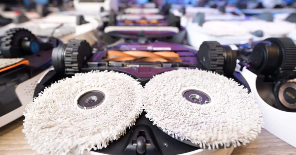 | Vacuum Wars Close-up of a robot vacuum’s underside showing two round, microfiber dual spinning mop pads mounted side by side, with other robot vacuums blurred in the background.