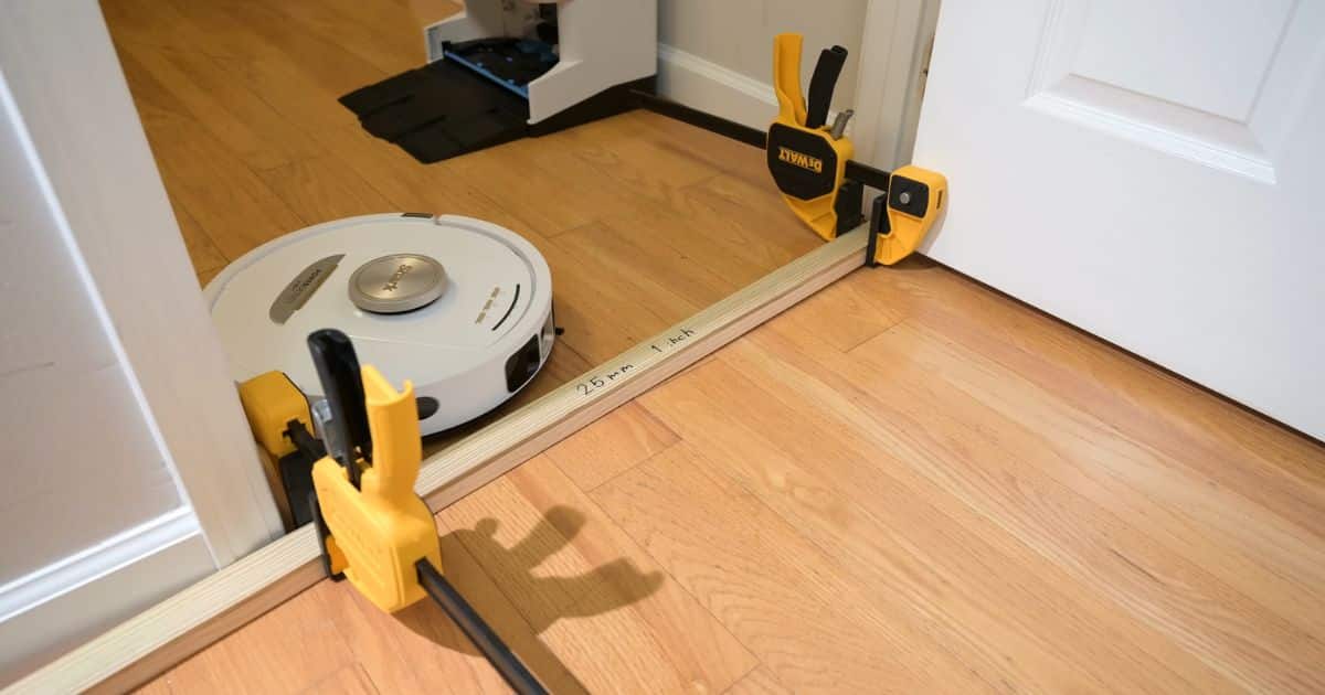 | Vacuum Wars Robot vacuum attempting a doorway threshold test with a wooden strip labeled โ25 mm / 1 inch,โ held in place by clamps; the robot stops short of crossing.