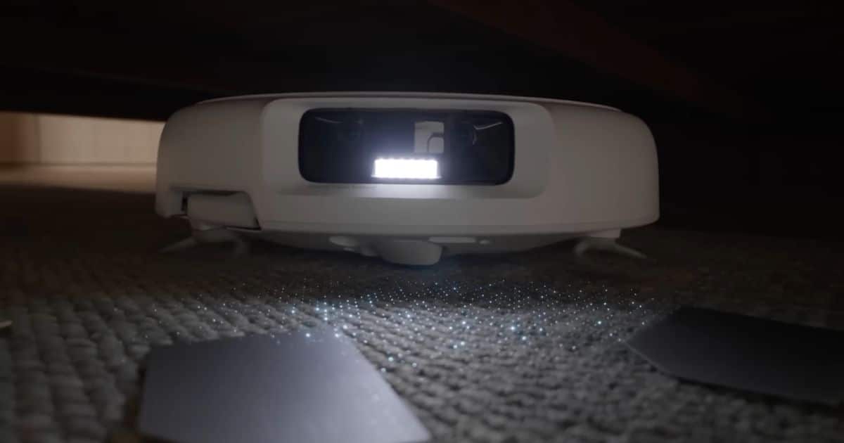 | Vacuum Wars DJI Romo robot vacuum under low furniture projecting a dotted light pattern from its front sensor window across carpet near small objects.