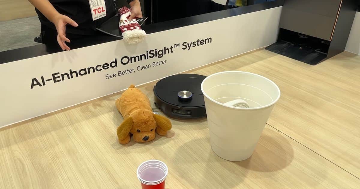 | Vacuum Wars Demo photo showing a robot vacuum on a show floor display with the sign “AI-Enhanced OmniSight™ System” and the tagline “See Better, Clean Better,” with props including a plush dog, a small cup of liquid, and a bucket near the robot.