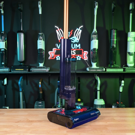Dyson Clean+Wash Hygiene in studio 450 ©Vacuum Wars | Vacuum Wars Cordless stick vacuum standing upright on a wooden table in a showroom, with other vacuums on display in the background.