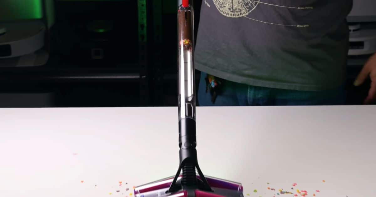 | Vacuum Wars Front view of the Dyson PencilVac standing on its Fluffycones cleaner head on a white test surface, with colorful cereal debris around the head and compacted debris visible high in the clear dustbin.
