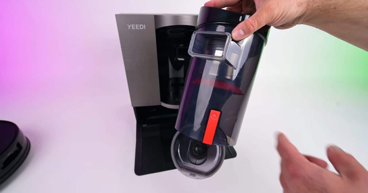 | Vacuum Wars A hand holds the removable clear dust bin from the Yeedi S20 Infinity bagless dock in front of the dock base.