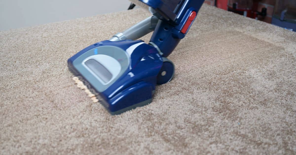 | Vacuum Wars Close-up of a blue Shark Navigator Lift-Away floor head on beige carpet pushing a line of cereal pieces forward, illustrating how the low front opening can snowplow larger debris.