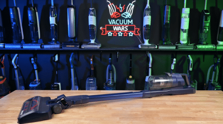 Bissell PowerClean DualBrush 4196K in studio 450 ©Vacuum Wars | Vacuum Wars Long vacuum wand on a wooden table, with a wall of upright vacuums behind and a 'Vacuum Wars' sign in the back.