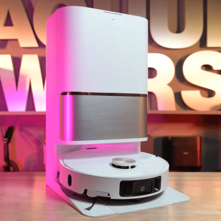Dreame L60 Ultra FE in studio 450 ©Vacuum Wars | Vacuum Wars White vertical smart‑home device with a circular robotic base on a desk, pink showroom lighting behind.