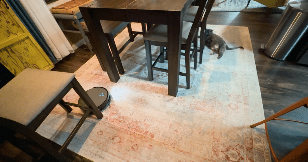 Ecovacs T90 Navigating at Home | Vacuum Wars The Ecovacs Deebot T90 Pro Omni cleaning around dining room furniture and rugs during a home test, with a cat nearby in the background.