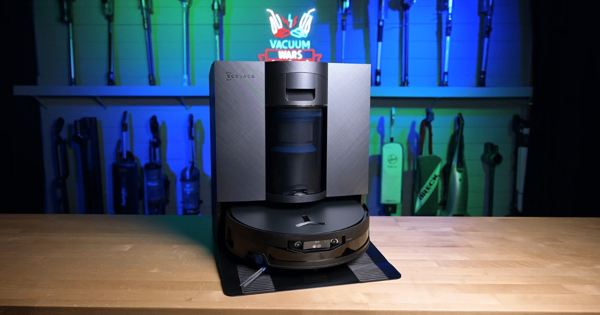Robotic vacuum on a wooden workbench in a tool-filled workshop, metallic body and docking base visible in front of blue-green lighting.