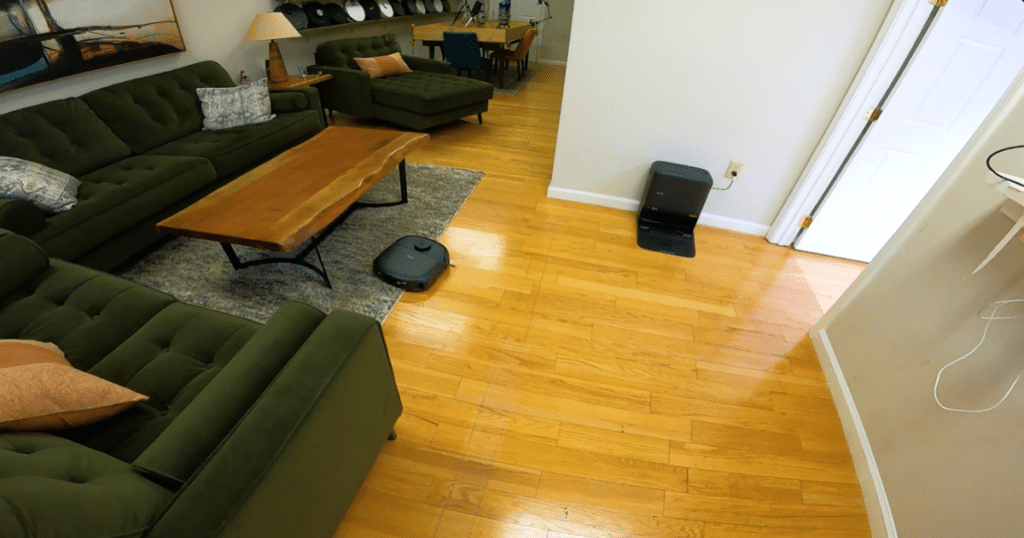 Eufy C28 hard floor and carpet cleaning | Vacuum Wars The Eufy Omni C28 robot vacuum cleaning a hardwood living room floor near its self-emptying multifunction dock station.