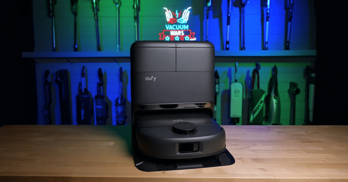 Eufy robotic vacuum cleaner on a wooden table, shown from the front in a workshop-style setting with blue-green lighting in the background.