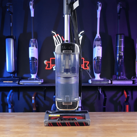 Canister vacuum cleaner centered on a wooden table with upright vacuums on display in a showroom poster.