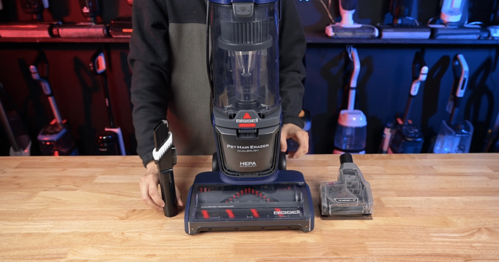 Bissell Pet Hair Eraser upright vacuum on a wooden table with a handheld brush and a crevice tool attached nearby, store display in background.