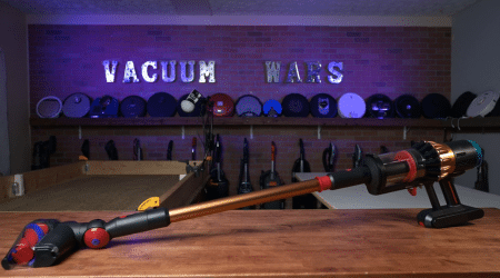 Dyson V16 Piston Animal in studio 450 ©Vacuum Wars | Vacuum Wars Long vacuum wand with attachments on a wooden table, behind it a row of canister vacuums on a brick shelf labeled 'VACUUM WARS'.