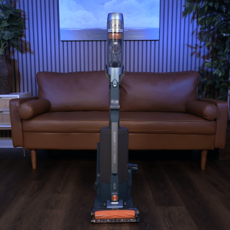 Shark PowerDetect Speed AutoEmpty IA3241 in studio 450 | Vacuum Wars Cordless stick vacuum standing upright in a living room in front of a brown leather sofa.