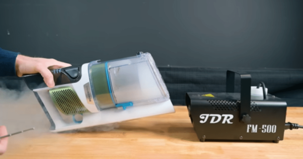 Shark PowerPro Flex Pet Filtration Test | Vacuum Wars Person operating a handheld fogging device connected to a black FM-500 unit, releasing mist onto a wooden table.