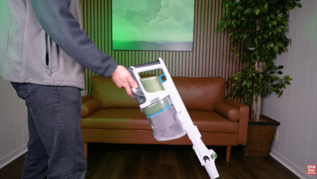 image | Vacuum Wars A person in a gray fleece jacket vacuuming a living room with a white cordless stick vacuum, brown sofa, and green plant in the background.