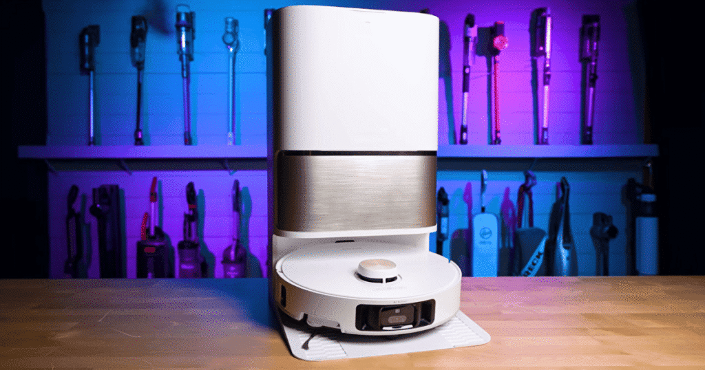 Dreame L60 Ultra PE hero | Vacuum Wars White robotic vacuum cleaner docked on its round charging base on a wooden workbench, with a wall of tools and colorful lighting in the background.