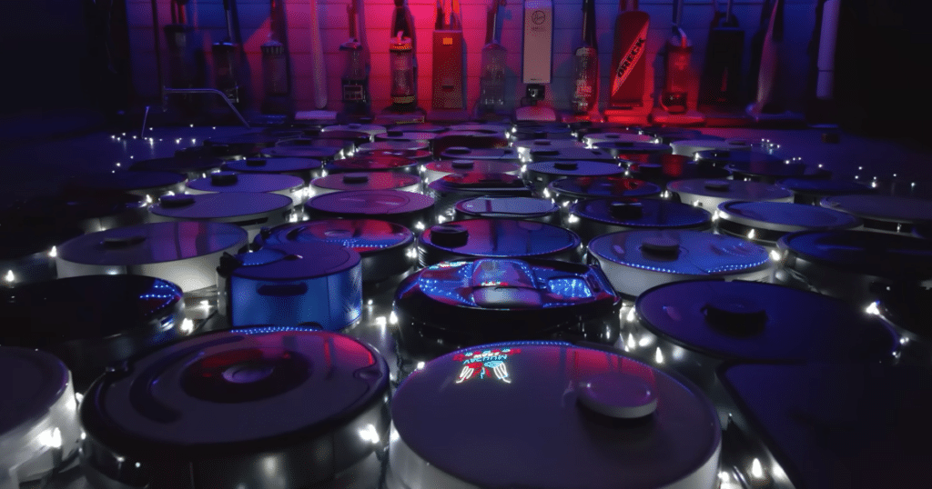Robot Vacuum Cleaners on Display at Vacuum Wars | Vacuum Wars A large collection of robot vacuums arranged across the Vacuum Wars testing studio under blue and red lighting.