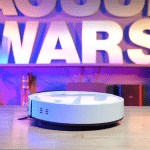 Ilife V2 in studio 150 | Vacuum Wars White robotic vacuum cleaner on a wooden floor with a Star Wars sign in purple-blue neon lights in the background.