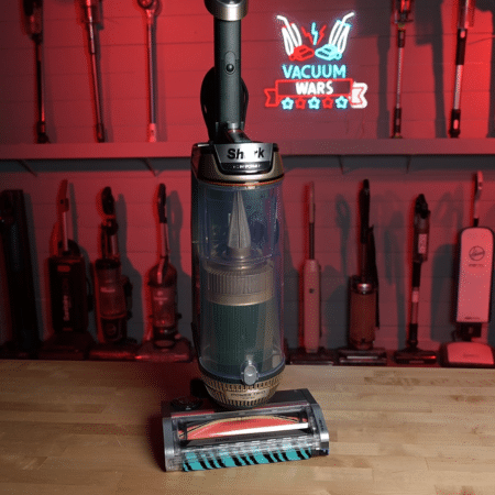 Shark PowerDetect Transformer in studio 450-2 | Vacuum Wars Upright Shark vacuum with a transparent canister on a wooden floor; red neon 'Vacuum Wars' sign and tool wall in the background.