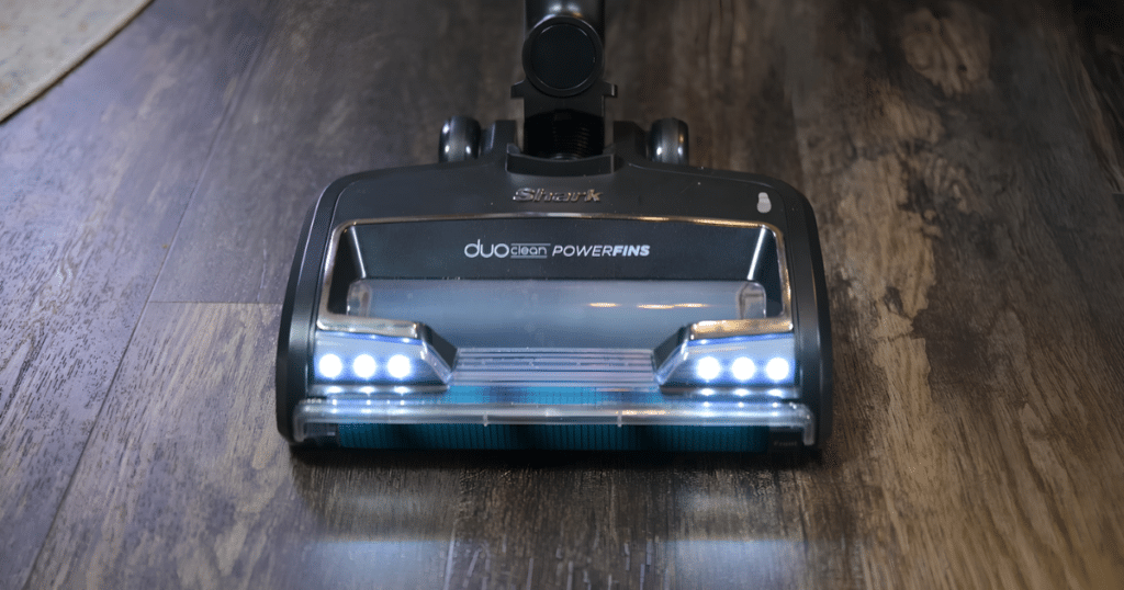 Shark Vertex Cordless Stick Vacuum DuoClean Powerfins Floorhead Vacuuming Hard Floor at Vacuum Wars | Vacuum Wars Shark DUOClean PowerFins vacuum head on a dark wooden floor with LED lights on.