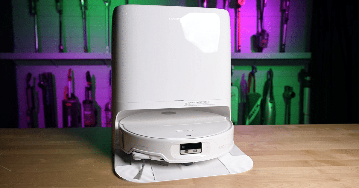 Roborock Qrevo Edge 3 Pro Robot Vacuum and Mop Combo docked in its tall charging station on a wooden table, with colorful vacuum cleaners visible in the background.