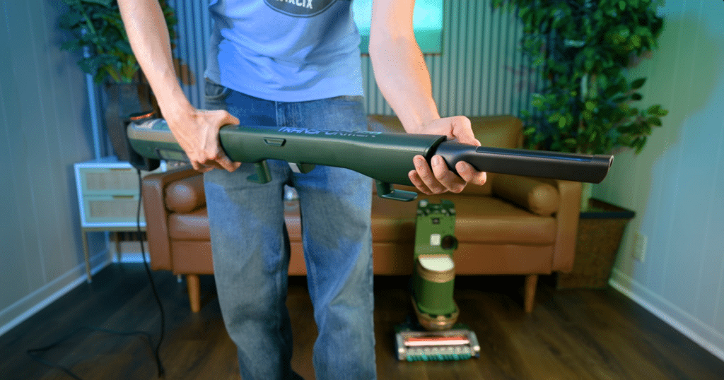 Shark Transformer PowerDetecthandheld stick with tool at Vacuum Wars | Vacuum Wars Person in a blue shirt holds a green wand-style vacuum cleaner in a living room, with a robotic vacuum on the floor behind them.