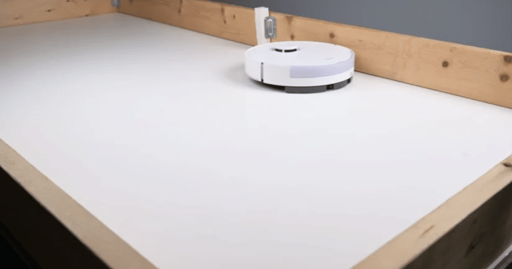 Dreame D20 Plus 7 Inch Hair Test 2 at Vacuum Wars | Vacuum Wars White robotic vacuum cleaner resting on a bright white floor in a corner with wood trim and an electrical outlet nearby during setup/cleaning.