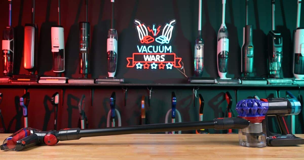 | Vacuum Wars Dyson V10 Konical cordless stick vacuum displayed horizontally on a wooden table in the Vacuum Wars testing studio, with other vacuums and an illuminated Vacuum Wars sign in the background.