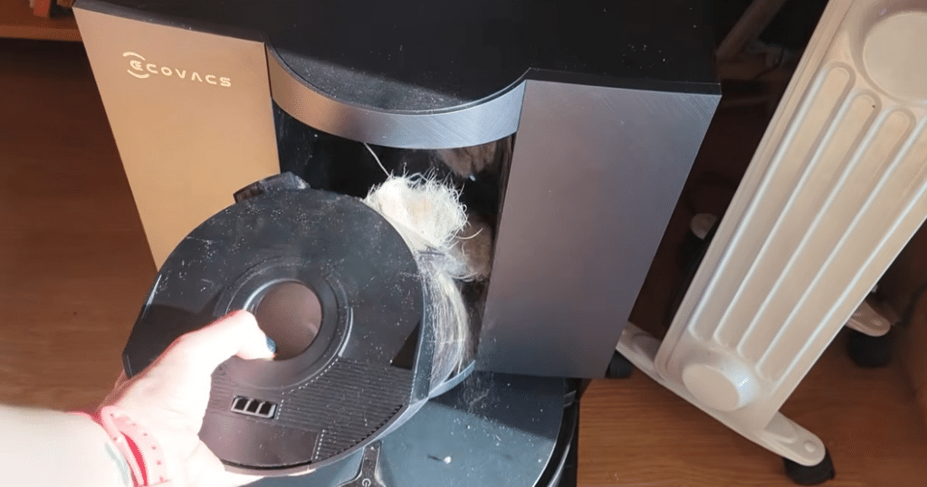 Ecovacs X12 Pet Hair Station Clog | Vacuum Wars Person removing the dust bin from an ECOVACS robotic vacuum, with hair tangled in the brush wheel area.