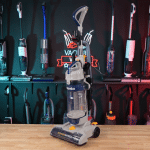 Eureka PowerSpeed E2 in studio 150 | Vacuum Wars Cordless stick vacuum with attachments on a wooden table, tools hanging on a display wall in the background.