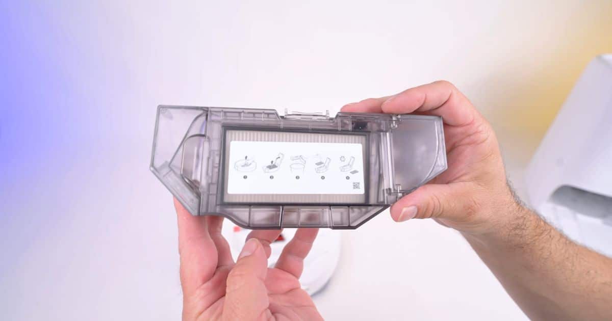 | Vacuum Wars Close-up of the Roborock Qrevo Edge 3 Pro's removable transparent dustbin being held in both hands, showing its internal filter and illustrated maintenance instructions.